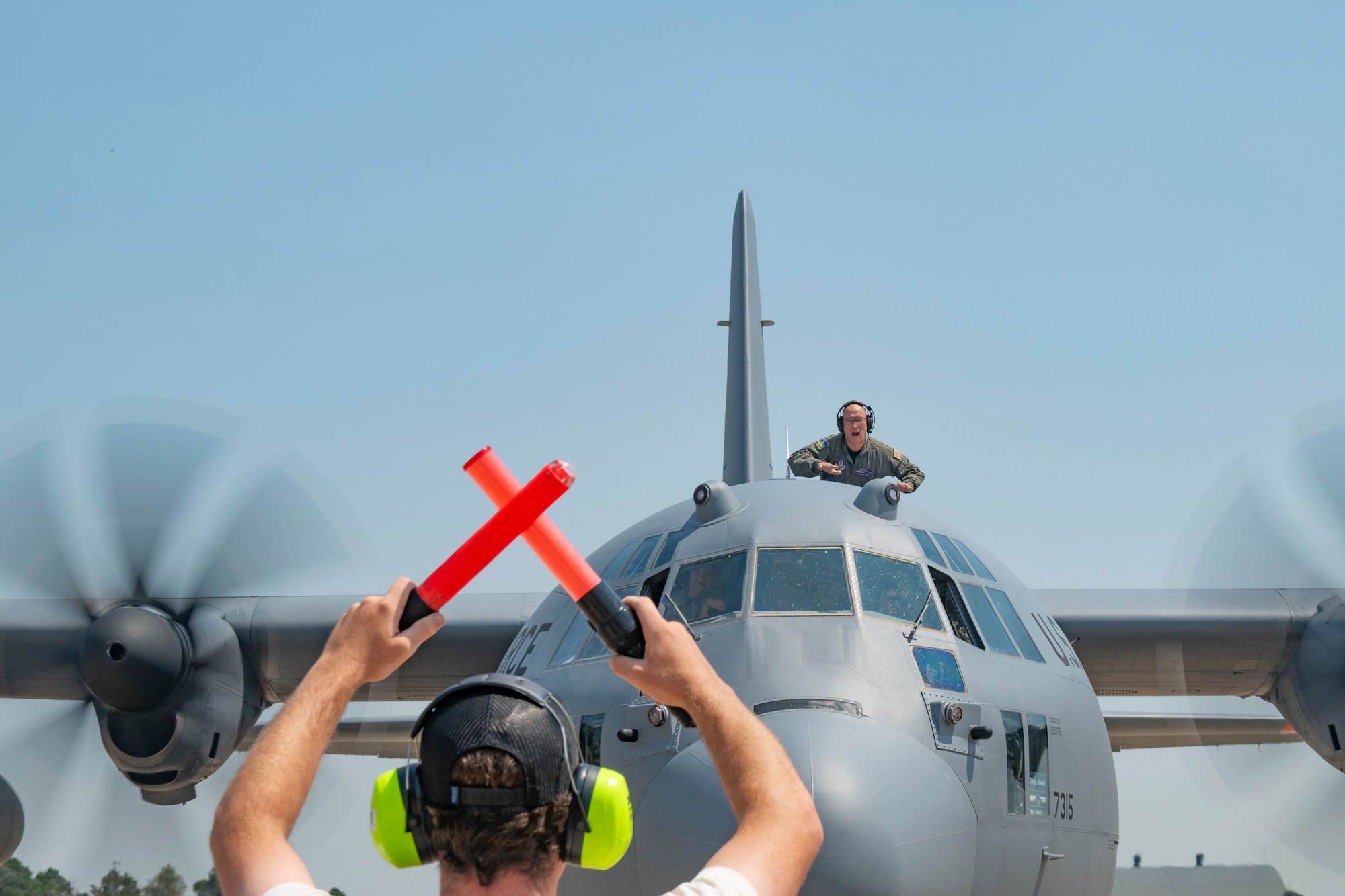 A man yells excitedly from a hole atop a military aircraft at another man holding aircraft marshalling wands.