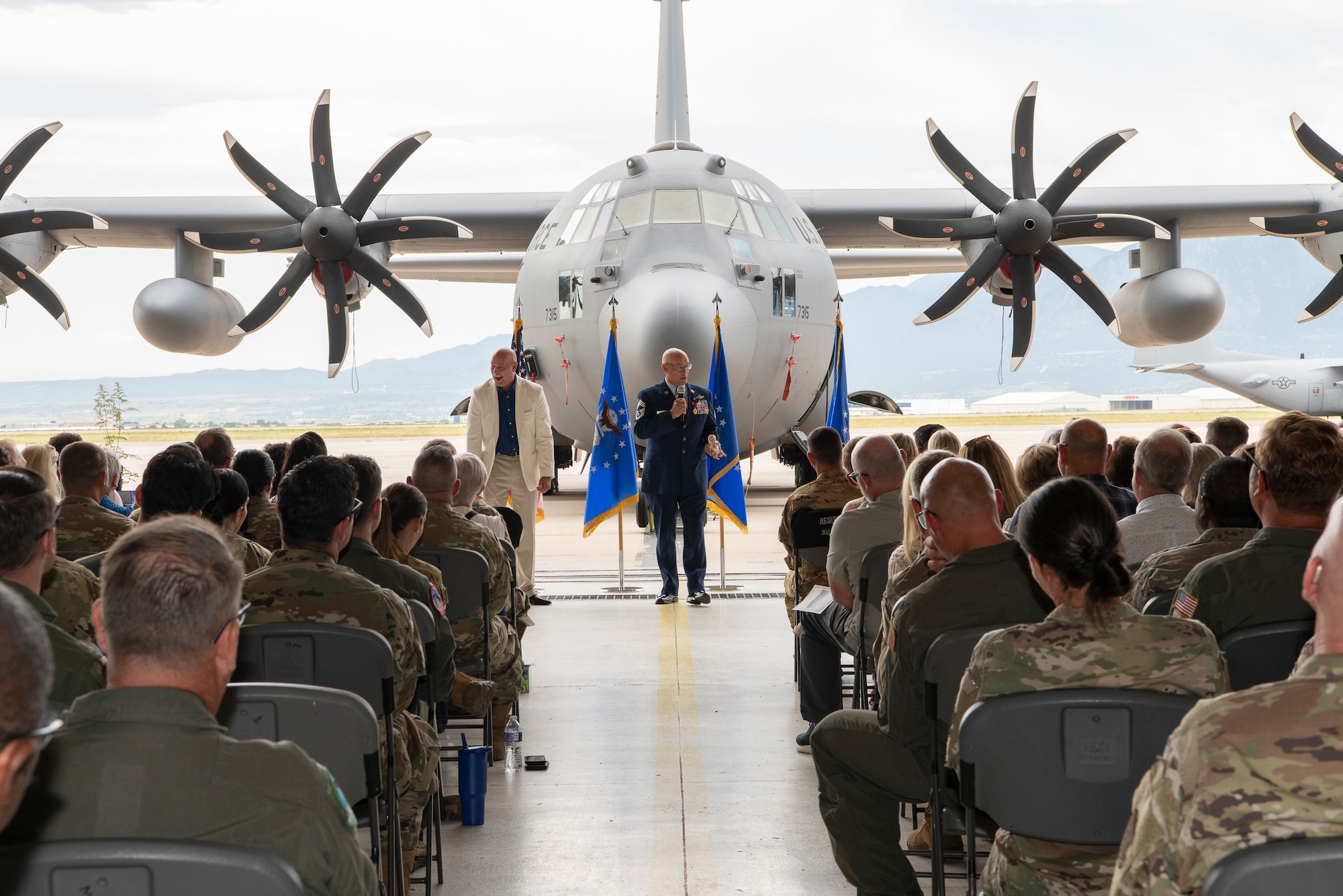 A man speaks to a crowd with a military aircraft in the background.
