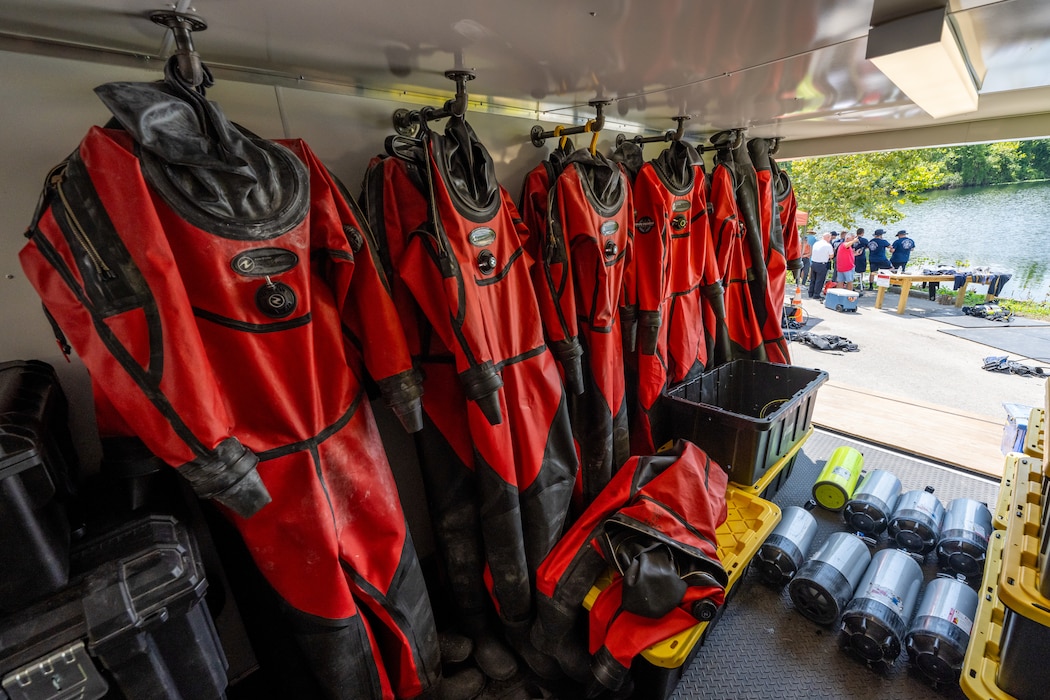 A row of several red and black scuba suits are hung along a white wall, eight oxygen tanks lay on the black floor. People standing outdoors near a lake front can be seen in the background