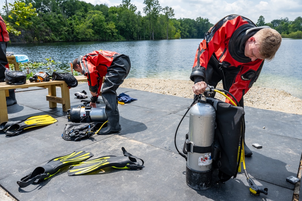 Two men, wearing red and black scuba suits, check their silver oxygen tanks by a lake front. Yellow and black flippers can be seen on the ground nearby
