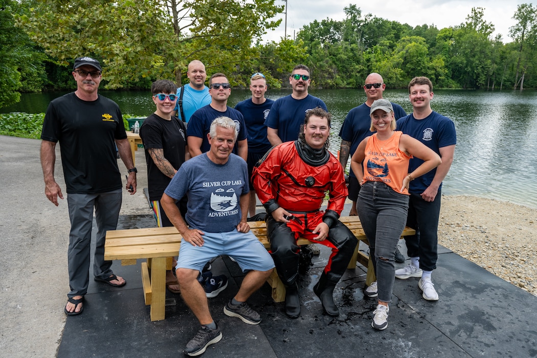 A group photo of several people, one man in bright red scuba dry suit front and center. A lake front is on the right side of frame, green foliage in the background.
