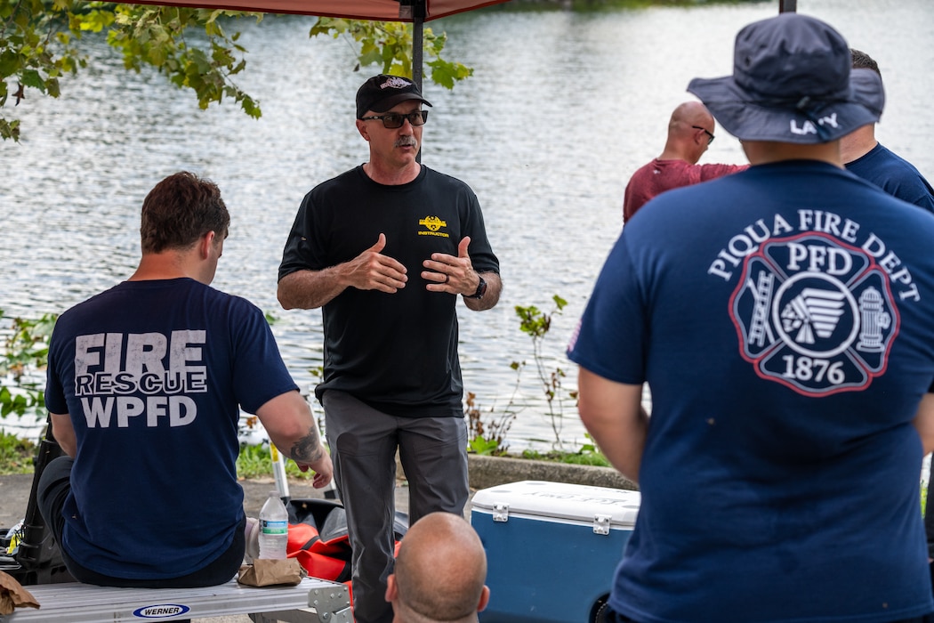 A man with a mustache and sunglasses, wearing a dark t-shirt and baseball cap, stands in front of a few other people with backs to viewer. Fire Dept logos can be seen on their backs