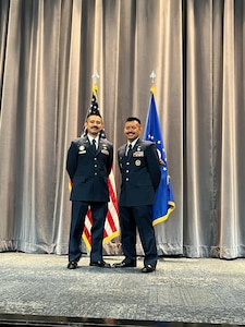 Two men stand on a stage in front of the U.S. flag and U.S. Air Force flag.