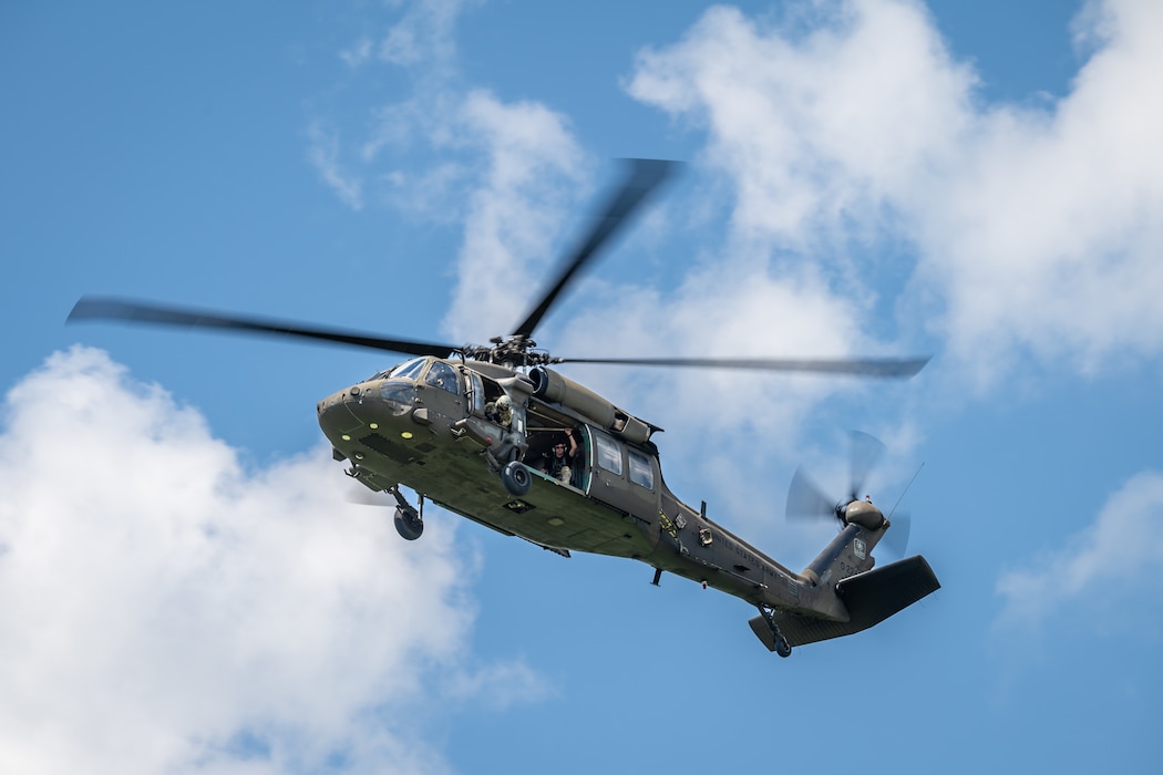 A dark grey helicopter flies in front of white clouds and deep blue sky