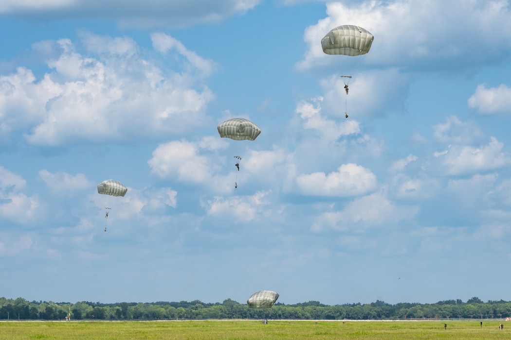 Three parachutes can be seen in the bright blue sky with staggered white clouds, one parachute at ground level.