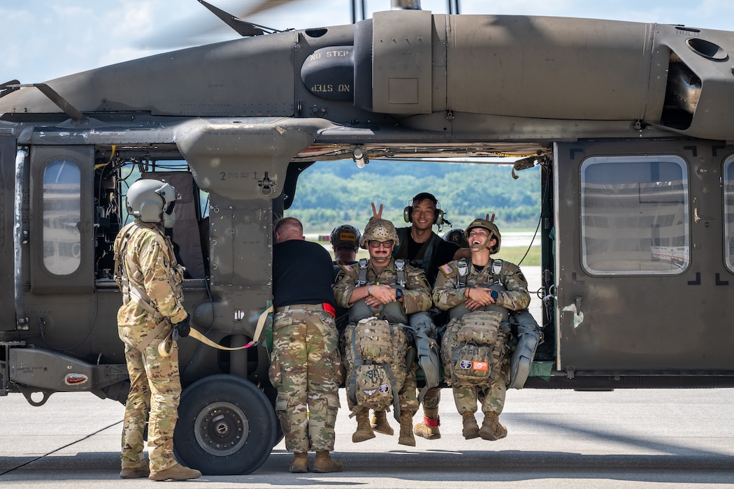 Two military members, wearing military camouflage uniforms, gear, and helmets, sit on the ledge of a helicopter frame as a man, wearing a black t-shirt, puts up "bunny ears" behind them. Other military personnel can be seen standing by the aircraft