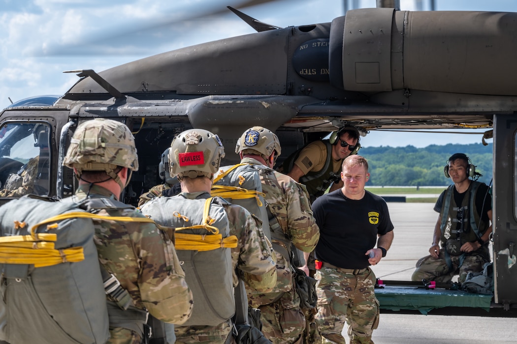 A shot from behind a single file line of Army soldiers wearing camouflage uniforms, helmets, and parachute packs leading up to the open bay of a military aircraft