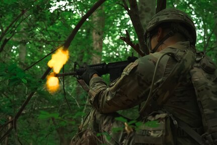 An Army soldier is firing blank rounds from a rifle in the woods, and there is an orange flash appearing at the muzzle.
