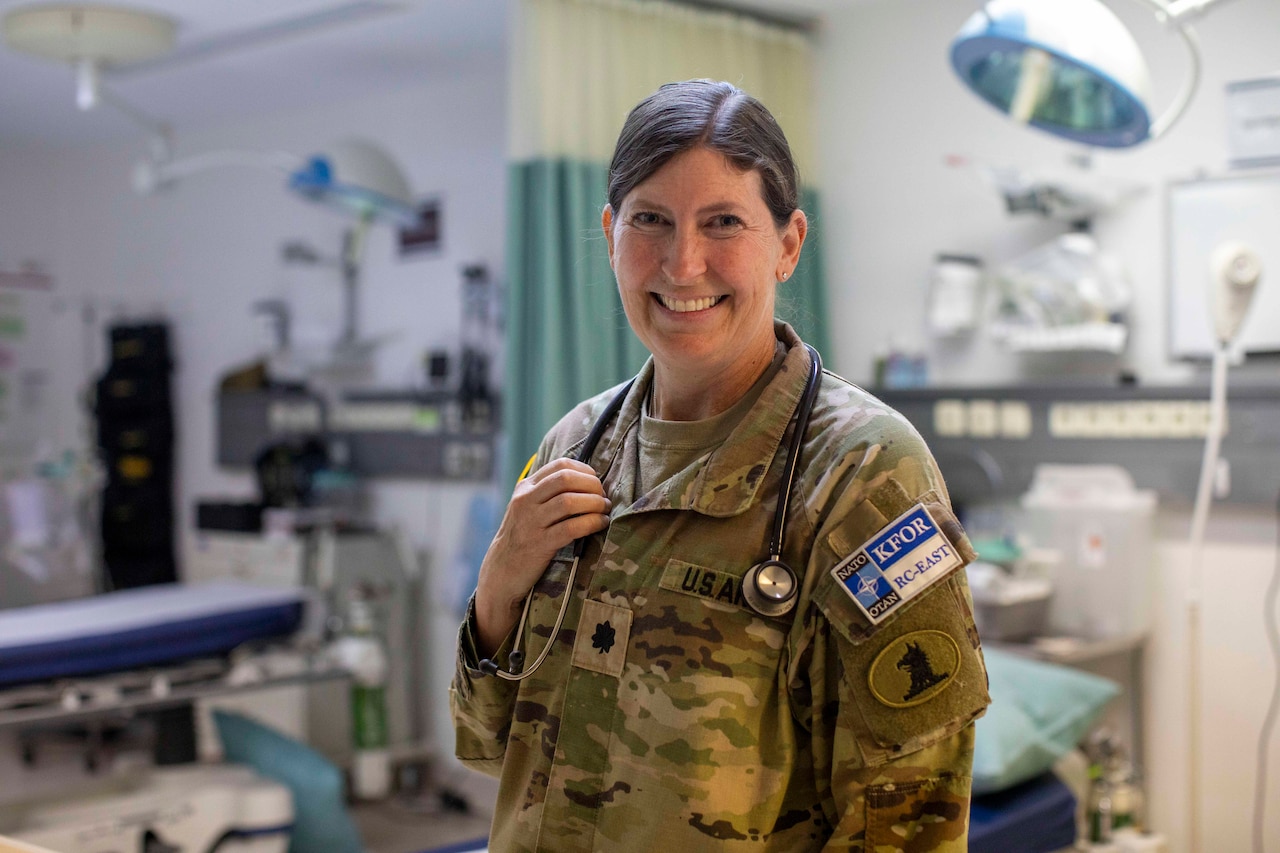 A woman dressed in a military camouflage uniform has a stethoscope around her neck and smiles as she poses for a photo in a medical treatment room.