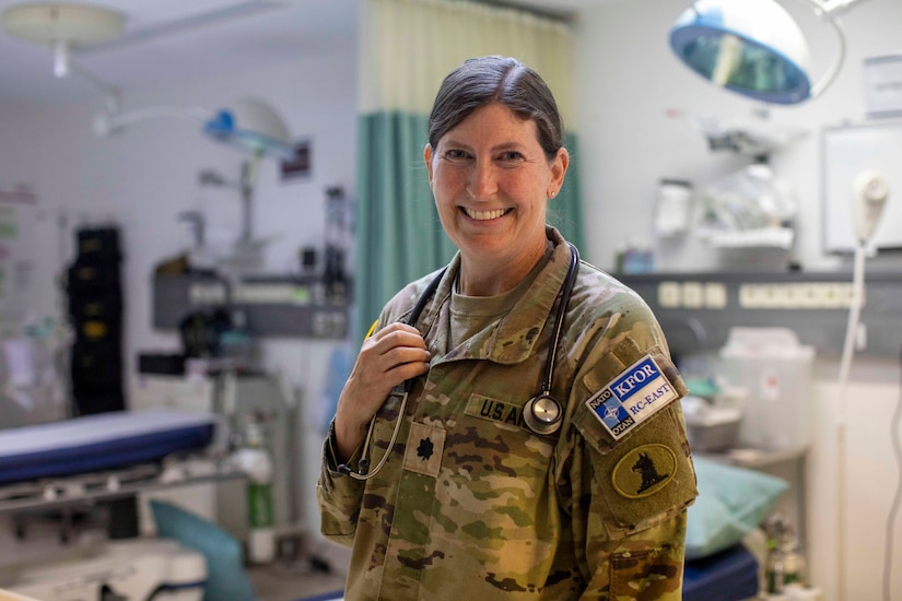 A woman dressed in a military camouflage uniform has a stethoscope around her neck and smiles as she poses for a photo in a medical treatment room.