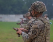 A row of Army soldiers in combat uniforms with helmets are adjusting their pistols at a firing range.