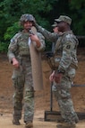 An Army soldier dressed in combat uniform is patting another similarly dressed soldier on the helmet as he walks by with a large target under his arm at a firing range.