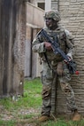 An Army soldier wearing combat uniform with helmet is holding a rifle across his chest while standing at the corner of a building.