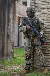 An Army soldier wearing combat uniform with helmet is holding a rifle across his chest while standing at the corner of a building.