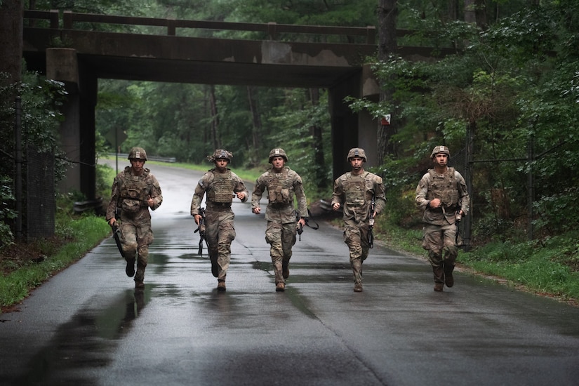Five Army Soldiers wearing combat uniforms with helmets are carrying rifles while running down a street just after an overpass.