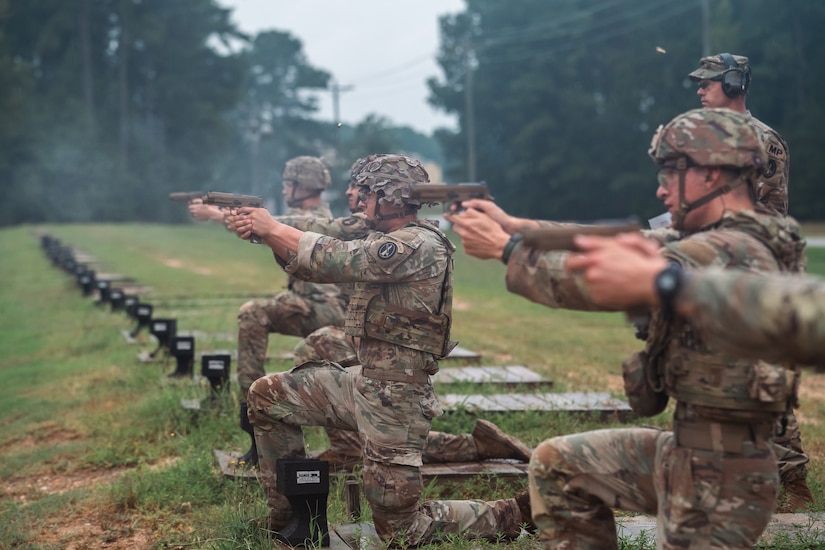 Army Soldiers in combat uniforms are kneeling on the ground in a row while firing pistols at a firing range.