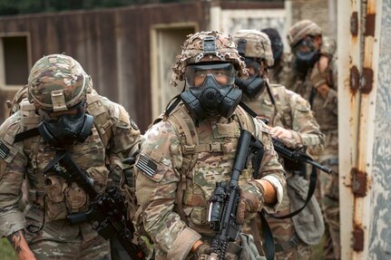 Army Soldiers wearing combat uniforms and gas masks are carrying rifles around a mock city environtment during an exercise.