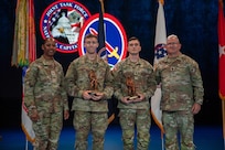 Four Army Soldiers wearing combat uniforms are standing in front of flags at a ceremony, and the two in the middle are holding large bronze trophies.