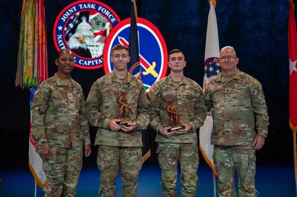 Four Army Soldiers wearing combat uniforms are standing in front of flags at a ceremony, and the two in the middle are holding large bronze trophies.