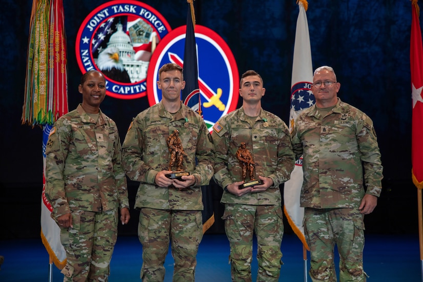 Four Army Soldiers wearing combat uniforms are standing in front of flags at a ceremony, and the two in the middle are holding large bronze trophies.
