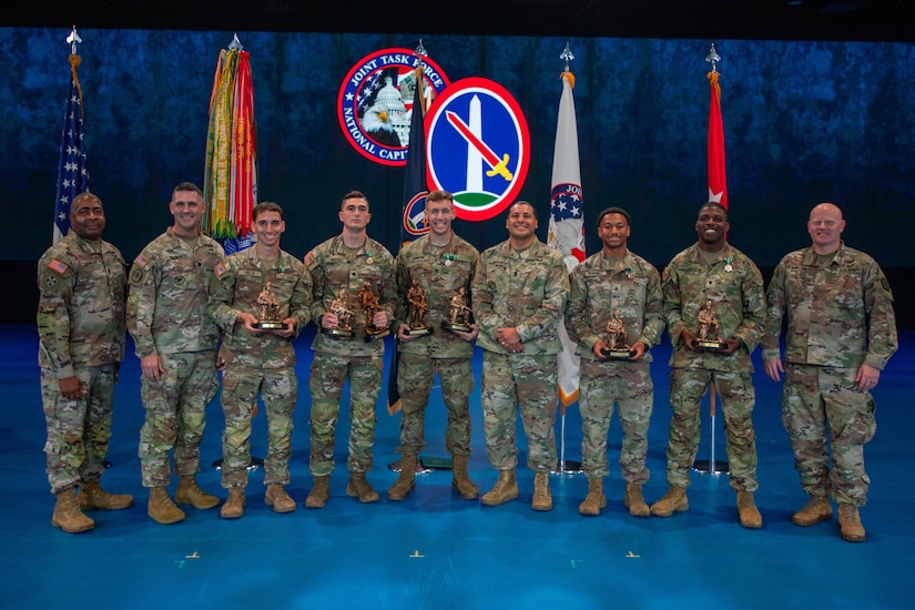 Five Army Soldiers are holding bronze trophies while posing for a picture with other Soldiers during an awards ceremony.