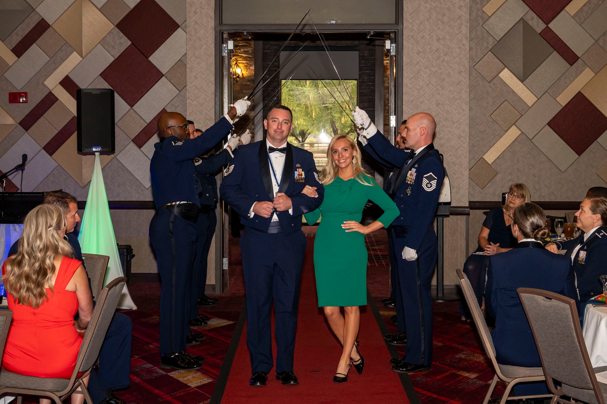 A newly inducted U.S. Air Force senior noncommissioned officer poses for a photo with his fiancée under a saber team arch during the SNCO induction ceremony in Tucson, Arizona, Aug. 15, 2025. The saber arch is a tradition symbolizing the inductee’s transition into the senior enlisted ranks with the support of family and fellow Airmen. (U.S. Air Force photo by Airman 1st Class Samantha Melecio)