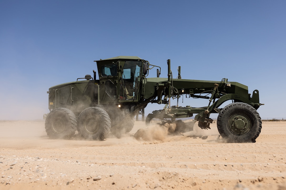 A maintenance vehicle kicks up dust while a Marine operates it on flat, desert-like terrain.