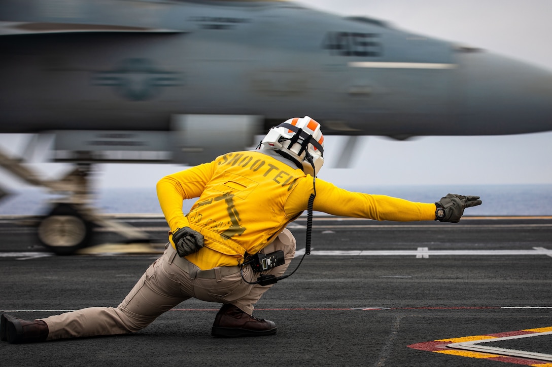 A sailor in yellow shirt, seen from behind, lunges and points to the right as an aircraft speeds in that direction on a ship's flight deck.