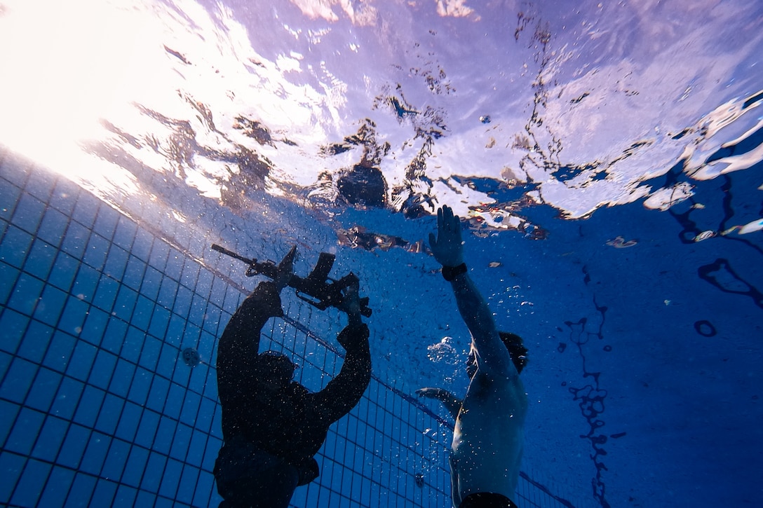 Two Marines stand submerged in a swimming pool, one in uniform and holding up a rifle.
