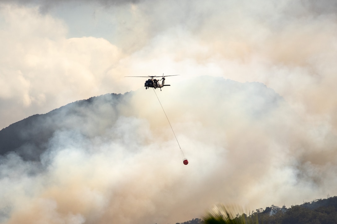 An Army helicopter with a bucket attached flies in a mountainous area in skies filled with smoke.