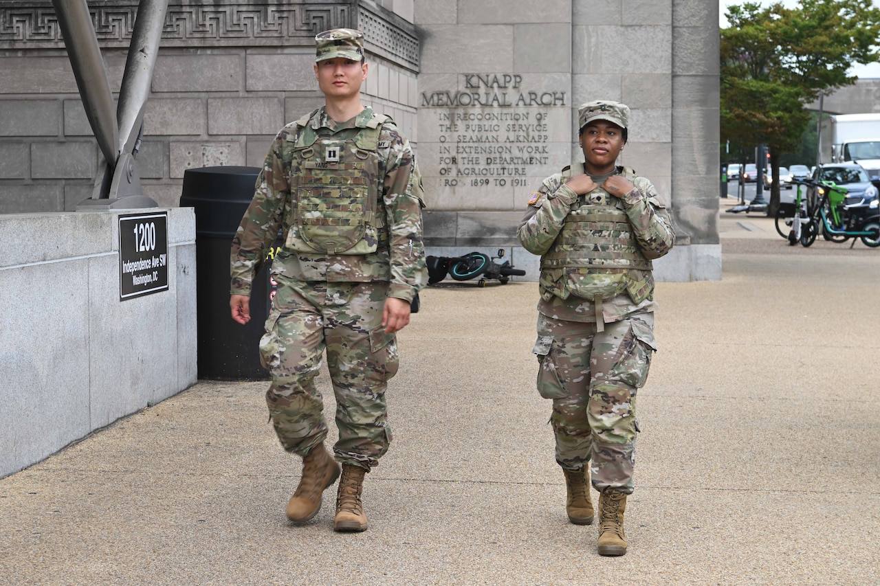 Two soldiers walk along a sidewalk, with a building and vehicles parked on the side of a street in the background.