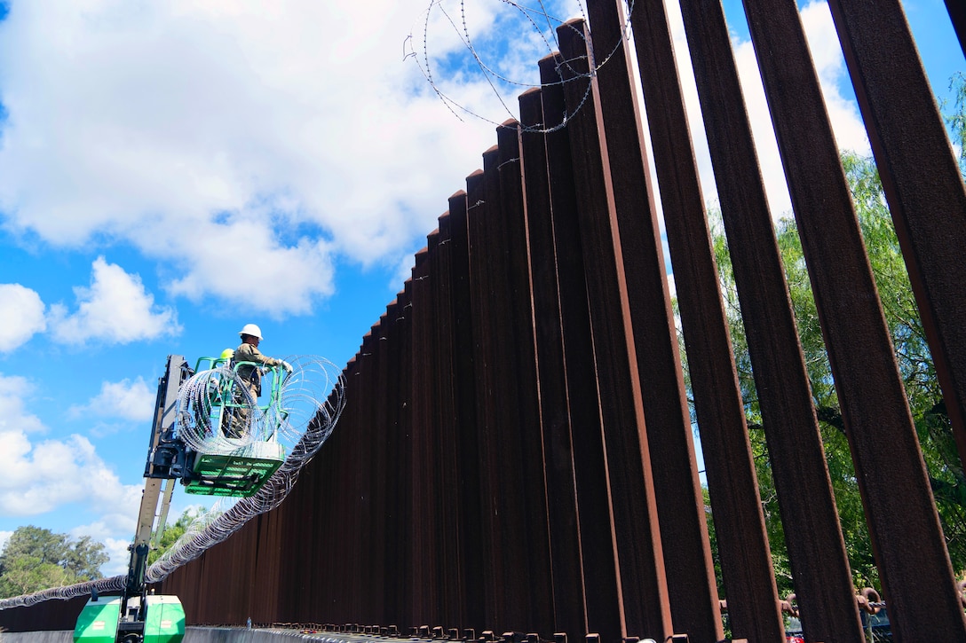 Two soldiers in helmets stand on a raised, green platform to install concertina wire on a tall, metal wall on a sunny day.