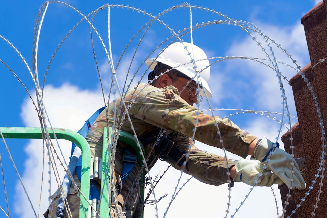 A guardsmen in a white helmet and gloves leans over a green platform to install concertina wire.