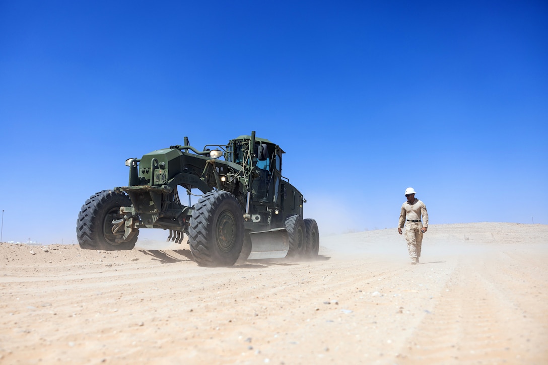 A Marine walks next to a dark green road maintenance vehicle driving on a sandy area under blue skies.