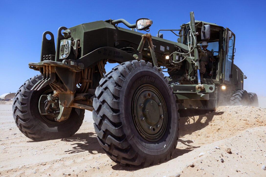 A Marine drives a dark green road maintenance vehicle on a dusty road under blue skies.