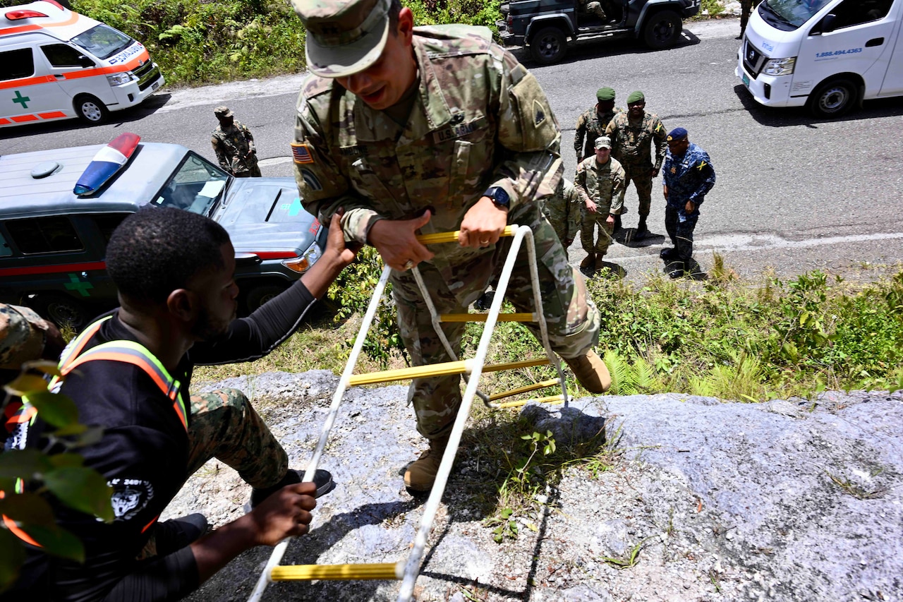 A man in a military camouflage uniform rappels down the side of a cliff while holding onto ropes. Another man dressed in a T-shirt and camouflage uniform pants watches and guides the man.