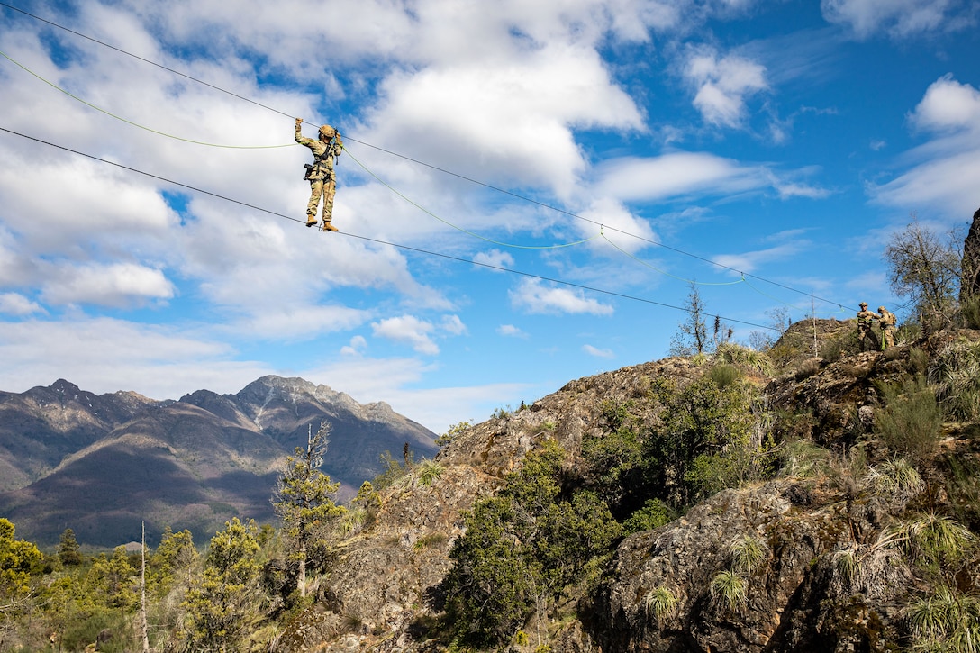 A soldier stands on a rope spanning a mountainous area while holding a second rope directly overhead.