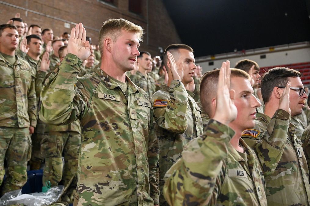 Dozens of soldiers stand on bleachers in a large room with their right hands raised.