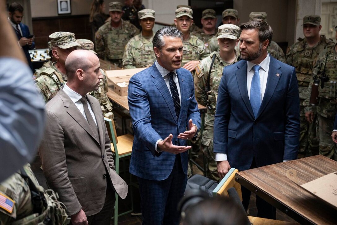 Three men wearing business attire talk to dozens of people in camouflage military uniforms.