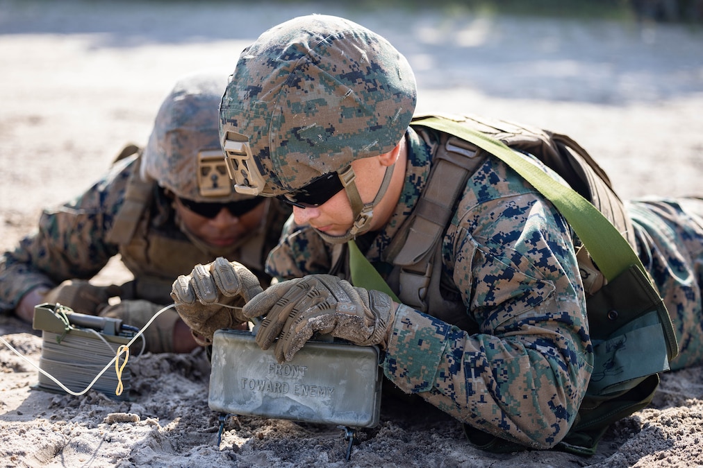 Two Marines lie on the ground as one connects wires to a rectangular mine.