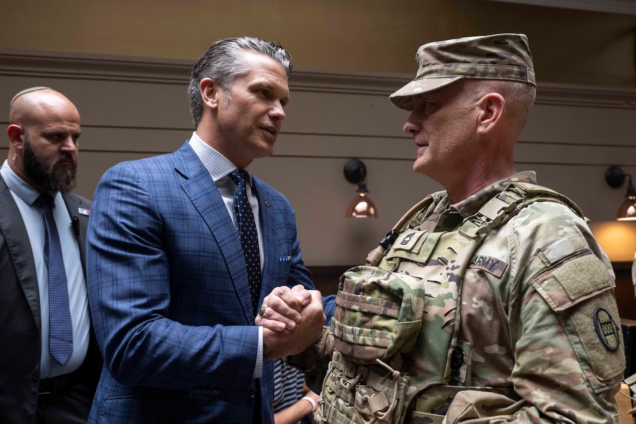 A man in a suit shakes hands with a service member in uniform inside a room as another man in a suit stands behind.