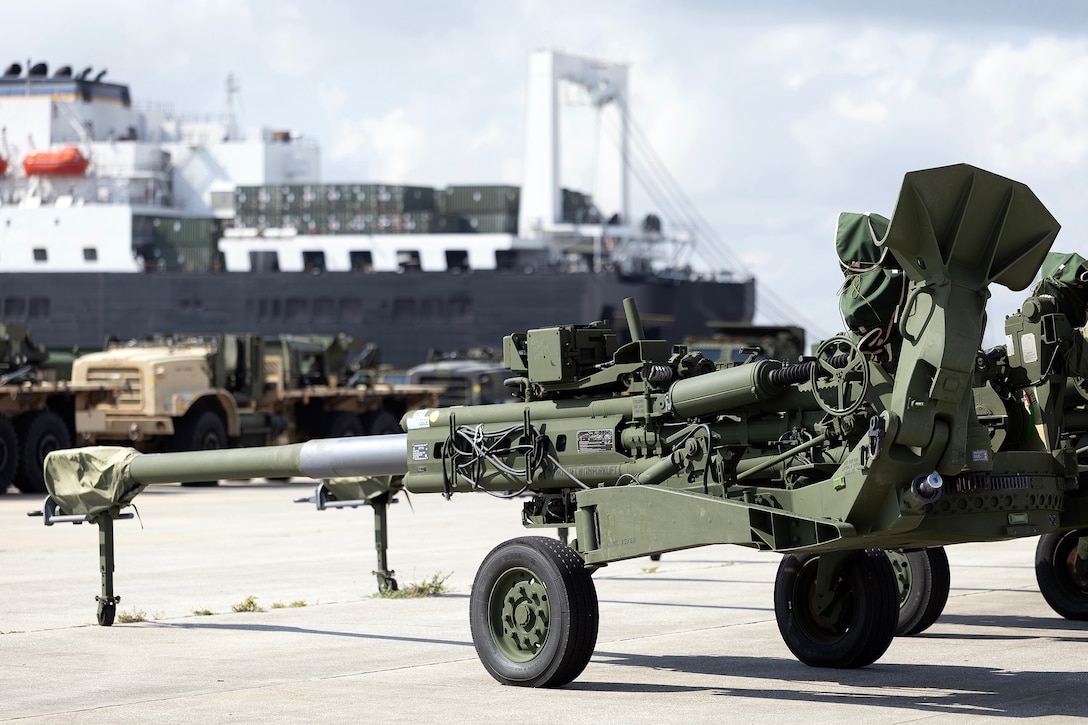 An M777 howitzer is staged Aug. 19, 2025, at Marine Corps Support Facility Blount Island in Florida, as the USNS Pililaau is backloaded nearby. The artillery pieces are returning to maritime prepositioning with a new recoil exerciser to preserve long-term readiness. (Official Marine Corps Photo/Dustin Senger)