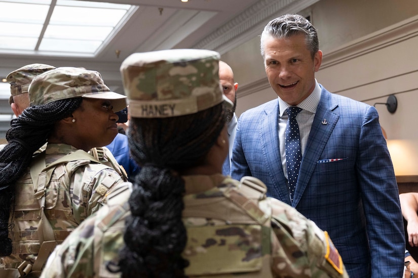 A man in a suit speaks with service members in uniform in a room.