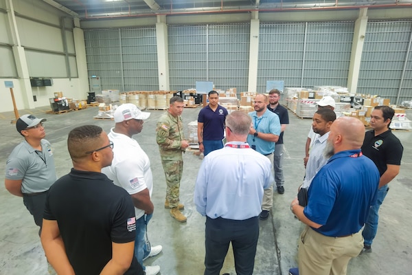 A man wearing a camouflage military uniform stands in a warehouse surrounded by a group of people.