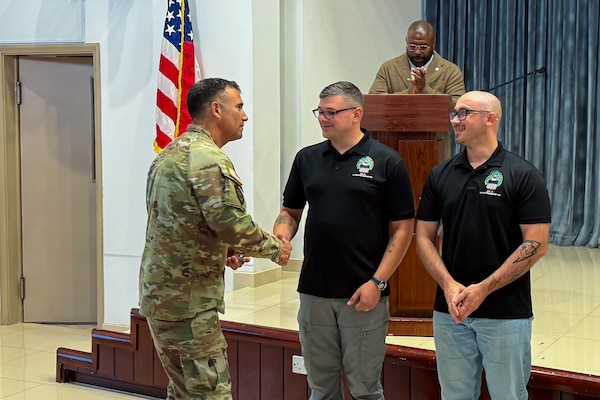 A man wearing a camouflage military uniform shakes a man's hand. There is another man standing next to them.