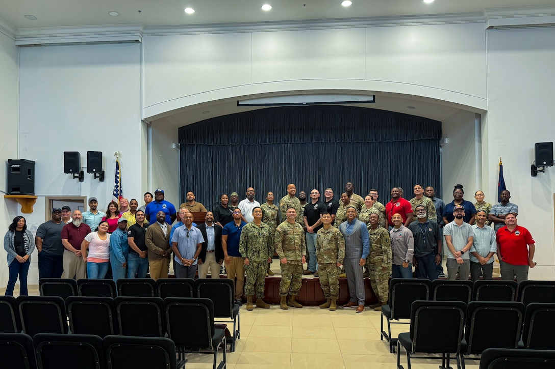 A group large group of people, four of them wearing camouflage military uniforms, stand in front of a stage posing for a group photo.