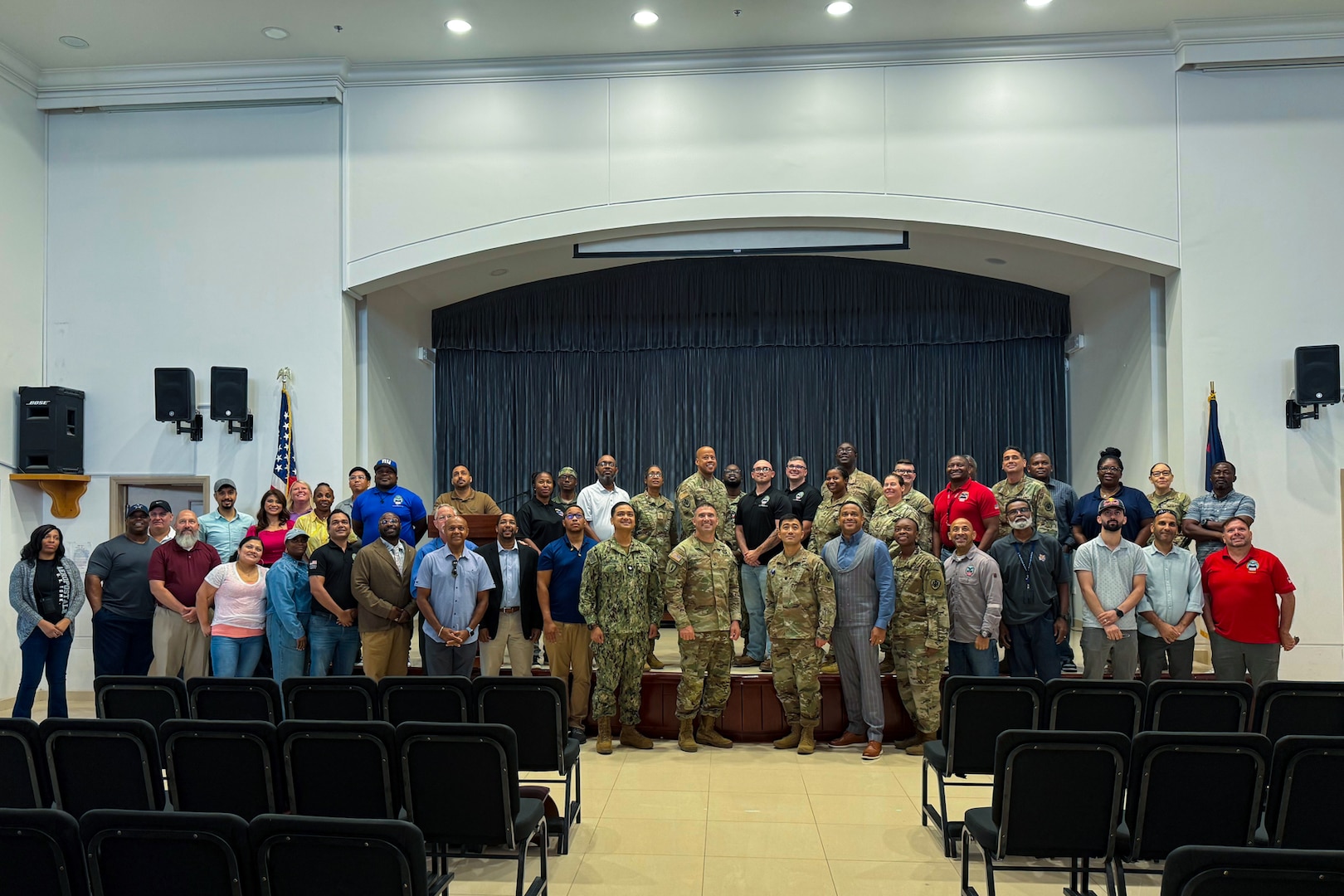 A group large group of people, four of them wearing camouflage military uniforms, stand in front of a stage posing for a group photo.