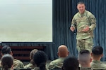 A man wearing a camouflage military uniform stands on a stage talking to an audience.