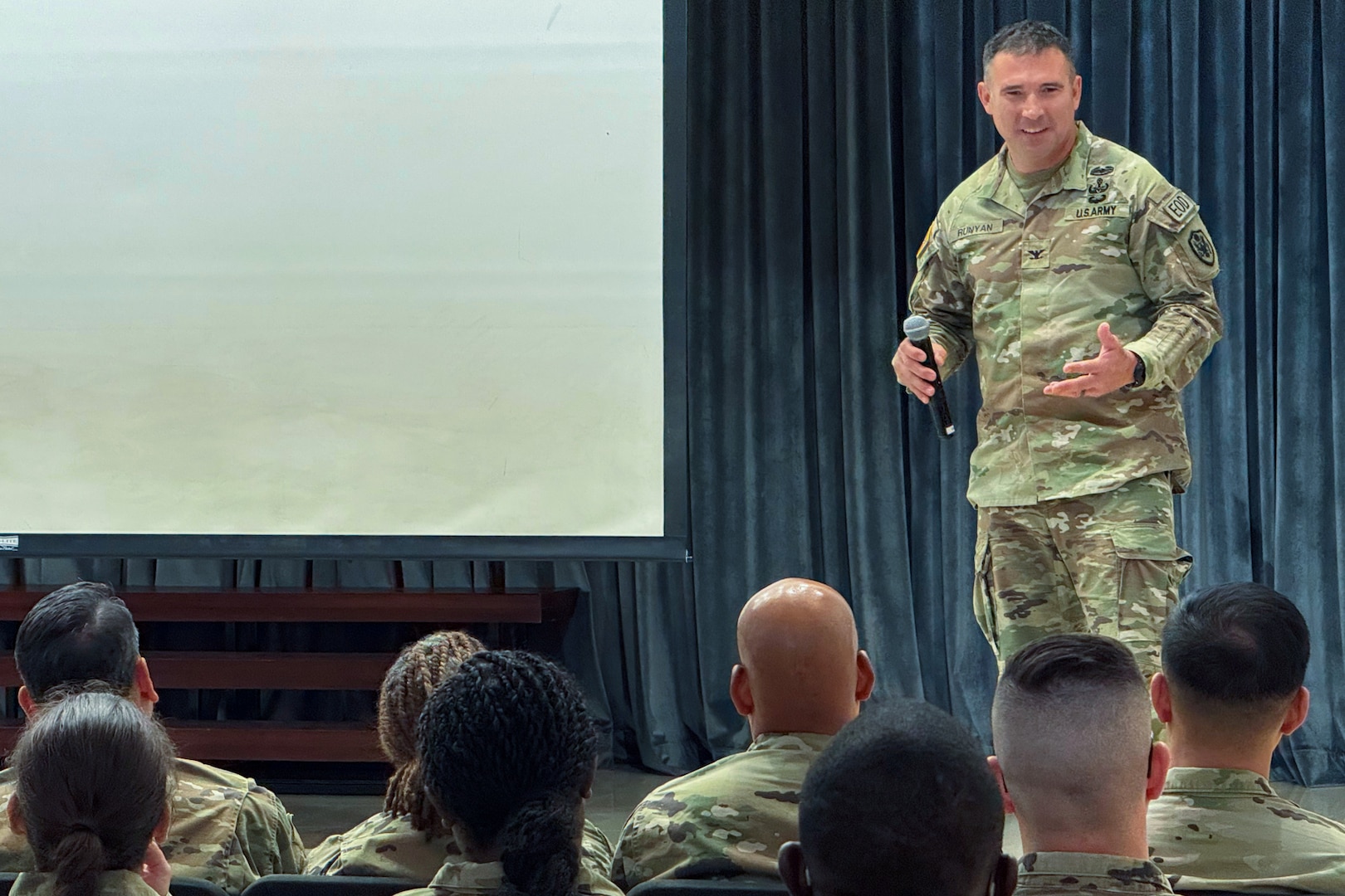 A man wearing a camouflage military uniform stands on a stage talking to an audience.