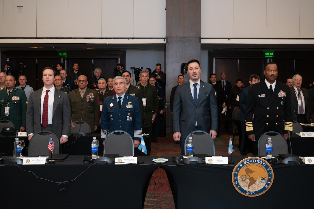 Two men in grey suits and two men in military uniforms stand in a line behind a table